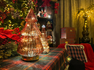 Decorative glass Christmas trees illuminated with warm lights are placed on a festive table adorned with plaid fabric, surrounded by holiday decorations and a cozy atmosphere