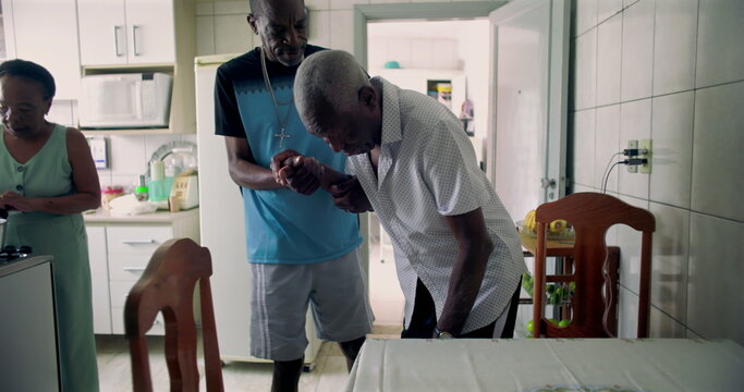 Elderly African American man being helped by his son to sit at a dining table in a cozy kitchen, reflecting care, compassion, and family connection in daily life - Powered by Adobe