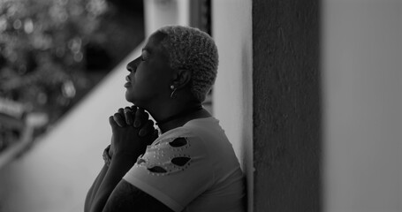 African American woman leaning against a wall, hands clasped in prayer, seeking divine guidance and strength during emotional turmoil, reflecting on life's challenges