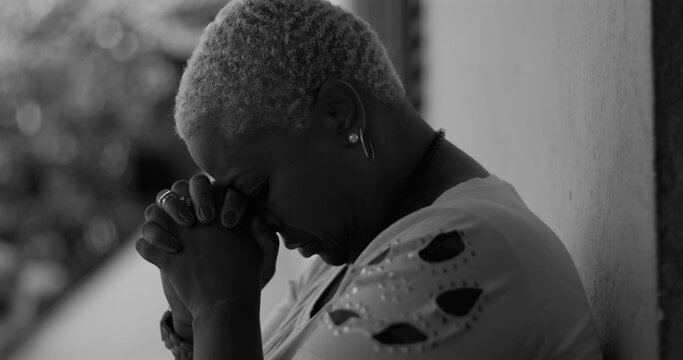 African American woman leaning against a wall, bowing her head in deep prayer, expressing emotional vulnerability and hope for strength amidst life’s adversities