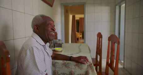 Elderly African American man smiling warmly while seated at a dining table in a cozy kitchen,...