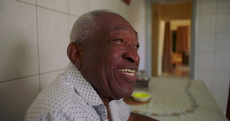 Elderly man laughing joyfully at dining table, vibrant smile and relaxed pose, sitting in kitchen with tiled walls, celebrating happiness and warmth in home setting