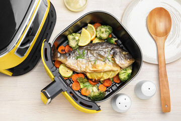 Cooked fish, lemon and vegetables in air fryer on white wooden table, flat lay