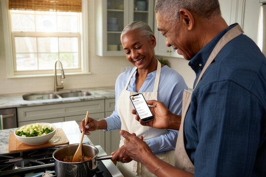 Elderly african american couple happily cooking in a bright kitchen, following a smartphone recipe - Powered by Adobe