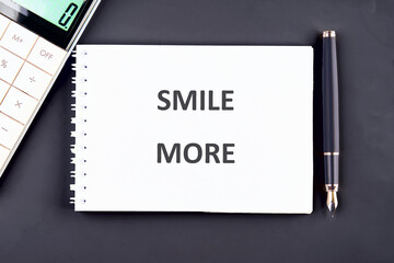 A clean workspace features a notepad with the encouraging phrase SMILE MORE clearly written. A calculator and a pen are nearby, creating a motivating atmosphere for productivity