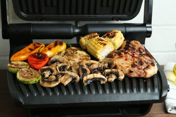 Electric grill with steak and vegetables on wooden table in kitchen, closeup