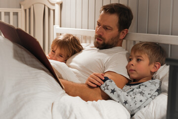 Fototapeta premium Happy mature 40 year old father reading book to his curious children lying in a bed before the sleep. Dad, little daughter and son spending time together. Family read the fairy story. Evening bedroom.