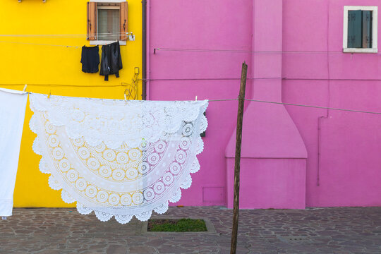 A large, intricately crocheted white tablecloth hangs on a clothesline, drying against vibrant pink and yellow house walls. A concept of traditional craftsmanship and daily life in a colorful village. - Powered by Adobe