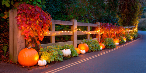 Autumn roadside decorated with orange and white pumpkins, glowing fairy lights, and red ivy on a wooden fence during dusk