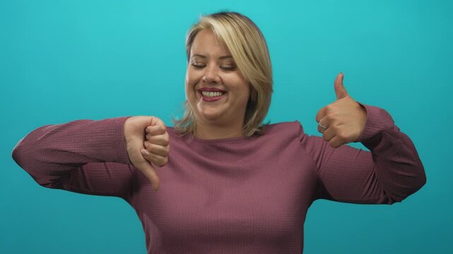 Young smiling woman shows dual bright thumbsup and thumbsdown gesture in turquoise studio; indecision.