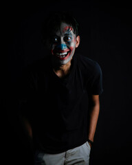 Grinning Young Man with Horror Clown Makeup and Sinister Expression. The dark background provides excellent copy space for a horror or event theme.