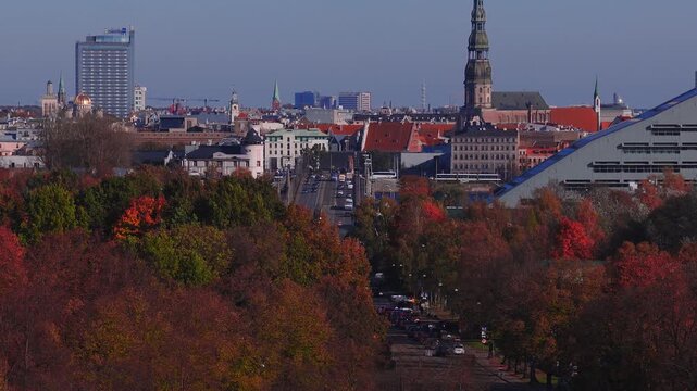 Aerial telephoto view moves over autumn trees toward central Riga. Cars and a tram travel a straight avenue. St. Peter's Church, the National Library of Latvia, Radisson Blu