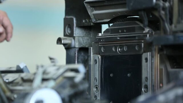 Skilled worker adjusts and maintains complex vintage printing press or typesetting machine. Detailed close-up of hands on intricate industrial components and levers.