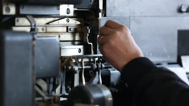 Skilled worker adjusts and maintains complex vintage printing press or typesetting machine. Detailed close-up of hands on intricate industrial components and levers.