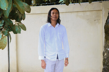 Serious Young Asian Man in White Shirt Standing Against Minimal Wall. He stands against a simple beige wall with leaves framing the top. Professional and minimalist style.
