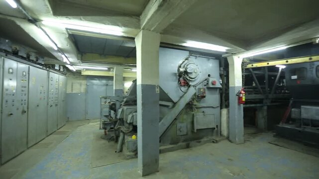 Interior view of an abandoned industrial bunker's machinery room. Features large mechanical equipment, concrete structures, and a long row of heavy electrical switchgear control panels.