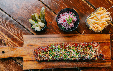 A long piece of meat is on a wooden cutting board with a side of pickles