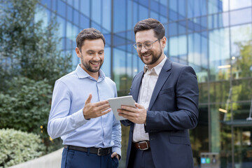 Two smiling businessmen looking at a digital tablet, discussing corporate strategy and working together outdoors in an urban setting with a modern glass building in the background