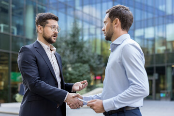 Two professional businessmen shaking hands in an outdoor setting, symbolizing a successful partnership, deal, or agreement after a corporate meeting in front of a modern office building