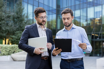 Two professional businessmen collaborating and analyzing documents, with one carrying a laptop,...