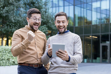 Two excited business colleagues celebrating triumph, looking at a digital tablet outdoors, standing...
