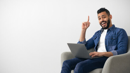 A young man sits comfortably in a gray chair with a laptop on his lap. He is smiling and pointing...