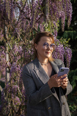 Woman Wisteria Smartphone - Smiling woman in a gray jacket holds a smartphone and stands near a...