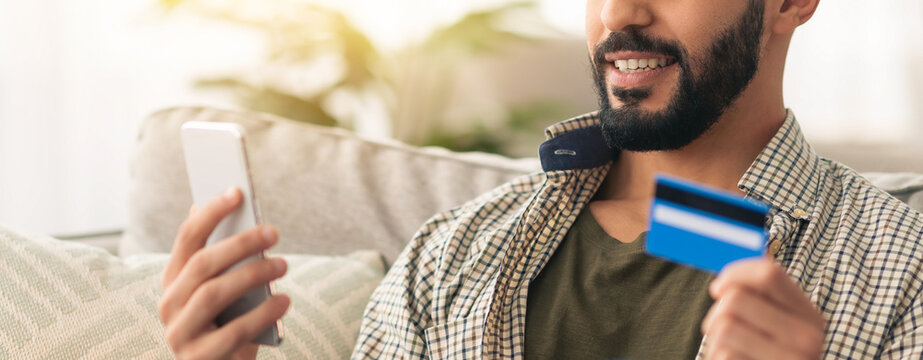 A man sits comfortably on a sofa, holding a smartphone in one hand and a credit card in the other. He appears focused and happy while shopping online in a bright living room.