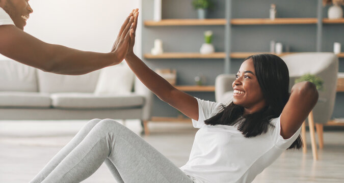 Happy laughing young black couple working on abdominal muscles at home, man holding female's legs doing crunches, giving high five in living room. Sport, fitness, exercising during lockdown quarantine