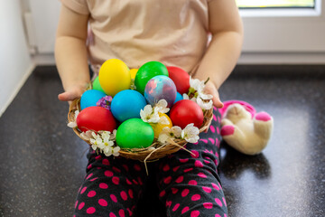 A little girl sits on a kitchen countertop and holds a set of colorful decorated Easter eggs in a wicker basket. Holy Easter