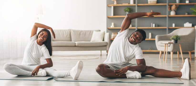 Millennial black couple doing lateral flexion exercise, working out together at home sitting on floor mat. Happy young lady and her boyfriend making side bending pose in living room looking at camera