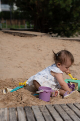 young child in a floral dress plays joyfully in a sandy playground with colorful toys, surrounded by wooden planks and greenery on a sunny day, joyful, peaceful, exploring, curiosity, sandbox