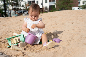 young child in a summer dress joyfully plays in the sandy playground, using colorful beach toys under bright sunshine, with a calm urban background of trees and buildings, playful, environment, joy