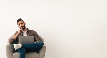 A man with a beard is seated in a cozy chair, looking thoughtful as he uses his laptop. The room...