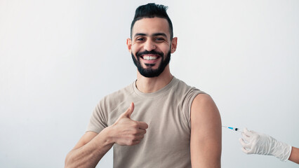 A young man shows a thumbs up while receiving a vaccination in a clean, well-lit clinic. He appears...