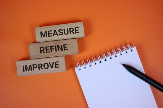 orange background with wooden blocks reading measure, refine, and improve beside a spiral notebook and black pen, symbolizing strategic planning, workflow optimization, and ongoing improvement.