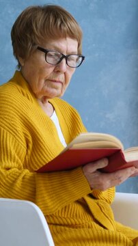 Focused senior woman with eyeglasses sitting in a chair and reading an interesting book with a red cover, enjoying her leisure time at home and cultivating her mind with a good story