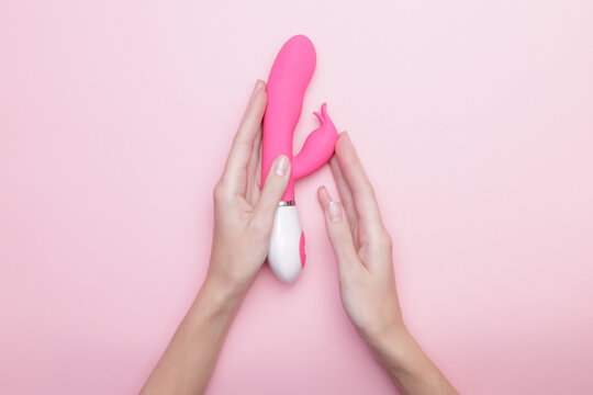 Female hands holding pink vibrator against soft pink background, top view