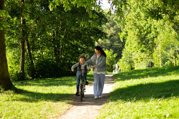 Happy cute boy learning ride bike in park on green meadow in summer day at sunset time. Parenthood and children holiday concepts.