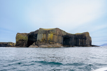 Wide view of Fingal’s Cave and the hexagonal basalt columns of Staffa Island, Scotland, with the ocean in the foreground and a pale blue sky.