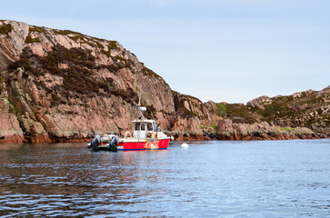 Small red and white fishing boat anchored in a sheltered rocky bay surrounded by rugged cliffs near the Isle of Mull, Scotland.