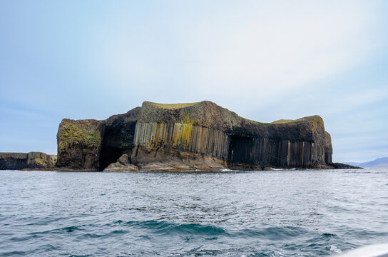 Wide view of Fingal&rsquo;s Cave and the hexagonal basalt columns of Staffa Island, Scotland, with the ocean in the foreground and a pale blue sky.