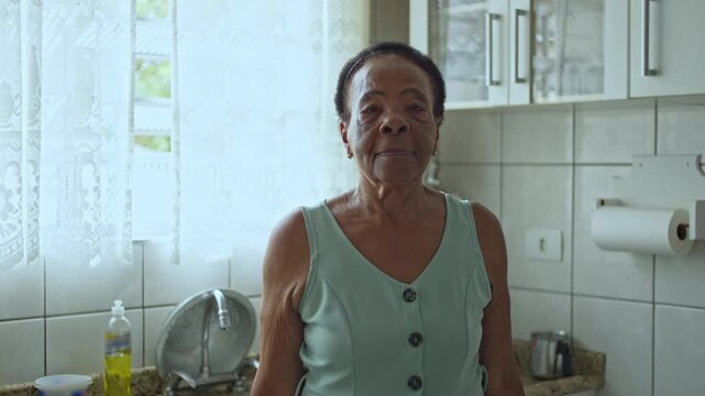 Calm Elderly Woman Staring At Camera While Standing In Bright Kitchen With Window Light Highlighting Her Facial Features