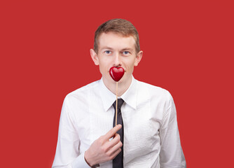 Man dressed in white shirt and black tie holds small red heart on stick near his chest, symbolizing...