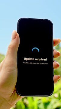 Vertical video. Smartphone in Hand Displaying Update Required Screen Against Sunny Sky and Green Grass Background, Concept Technology Maintenance and System Refresh