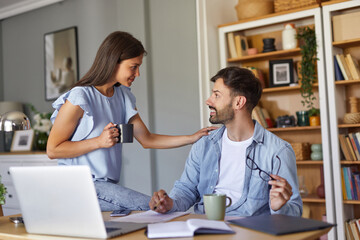 Fototapeta premium A man and woman interact warmly in their home office. The woman stands next to the man, who is seated, both enjoying coffee and sharing smiles while surrounded by books and a laptop.