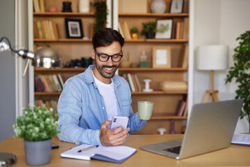 A man working in a cozy home office setting, checking his smartphone while sipping a beverage and surrounded by bookshelves, plants, and a laptop on the desk.