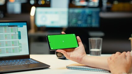Businesswoman looking at green screen on mobile phone during late night work session, finishing management tasks after hours in corporate office space. Using isolated copy space. Camera B.
