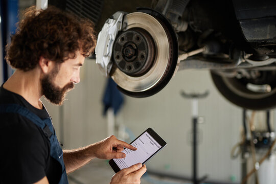 A car mechanic inspects service information on a tablet in a busy workshop. The vehicle is lifted, exposing its wheel and undercarriage for repair tasks.