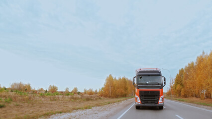 Fototapeta premium Red truck with goods in cargo trailer driving down a multi-lane road through a wooded area. International cargo delivery. Autumn landscape.